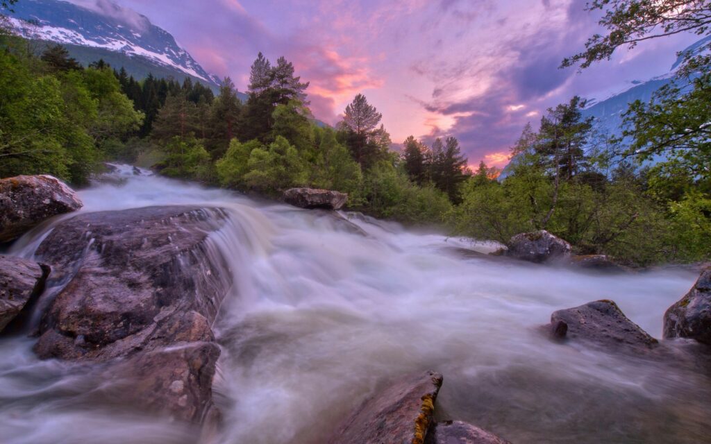 Sunset river flowing over rocks with lush green trees and snowy mountains in vibrant landscape Wallpaper