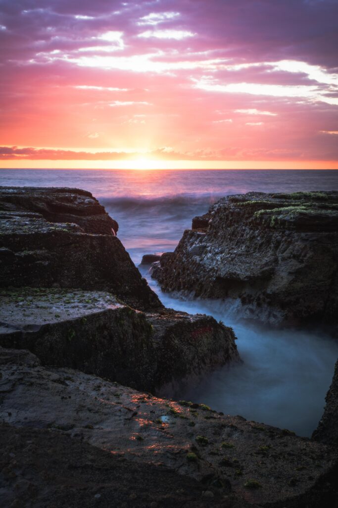 Breathtaking sunset over sea with vivid orange pink purple sky and waves crashing on rugged rocks Wallpaper