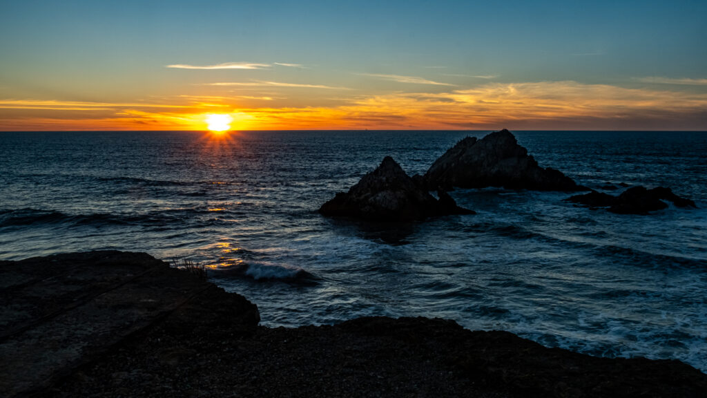 Glowing sunset over tranquil dark waters with jagged rocks silhouetted in vibrant seascape wallpaper
