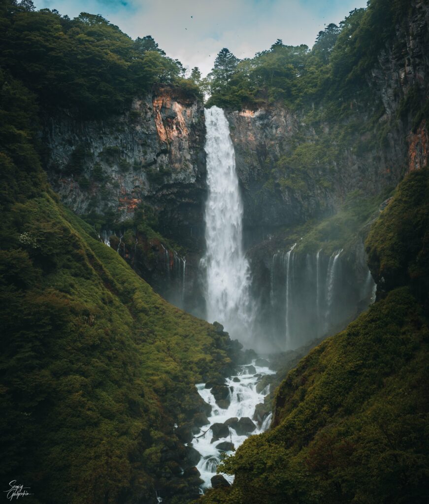 Powerful waterfall cascading down rugged cliff surrounded by lush green trees and mist background Wallpaper