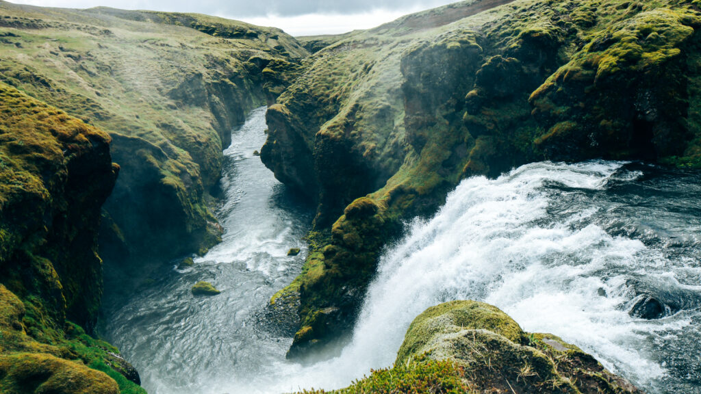 Waterfall cascading over mossy rocks into river surrounded by lush green hills and cliffs wallpaper