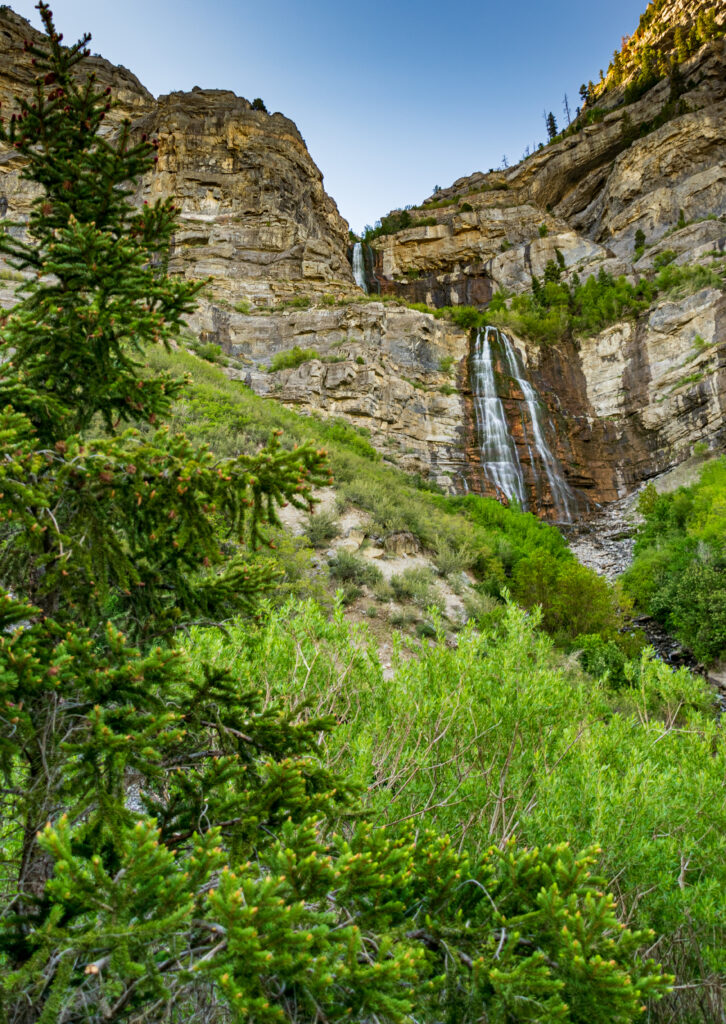 Towering rocky cliffs with lush green trees and a cascading waterfall under clear blue sky wallpaper