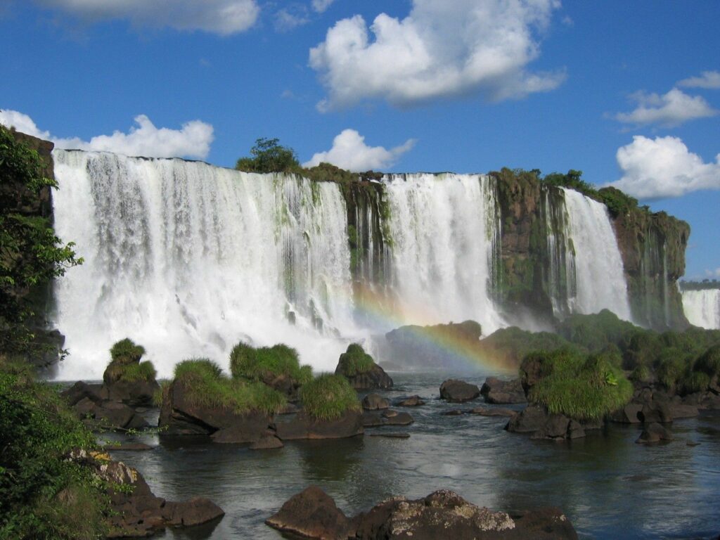 Vibrant waterfall over rugged rocks with lush greenery and rainbow under blue sky wallpaper background