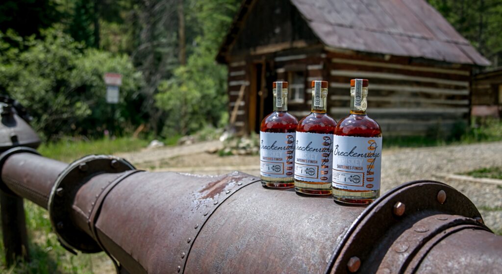 Breckenridge Distillery Sauternes Finish Whiskey bottles on rustic pipe with forest cabin backdrop Wallpaper
