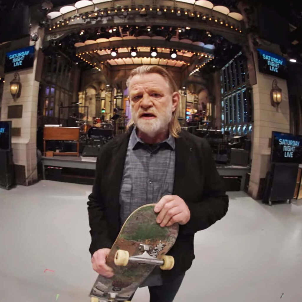 Brendan Gleeson on Saturday Night Live stage holding skateboard brightly lit dynamic background Wallpaper