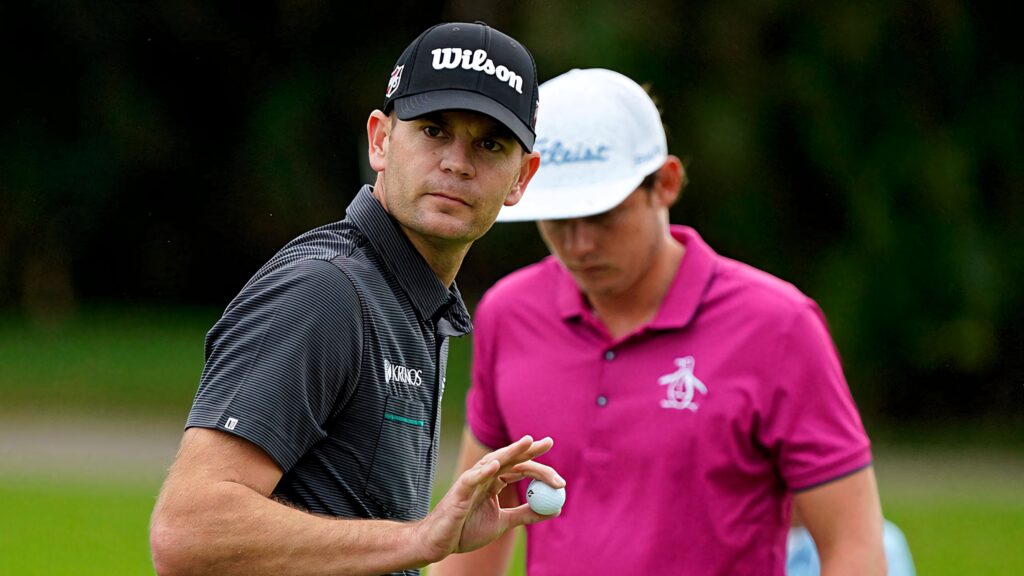 Brendan Steele holding golf ball gesturing on green course wearing Wilson cap with golfer nearby Wallpaper