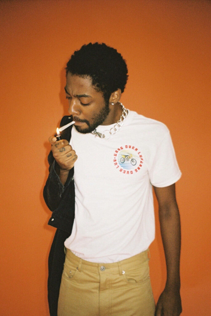Brent Faiyaz lighting cigarette vibrant orange background white tee tan pants chain necklace wallpaper