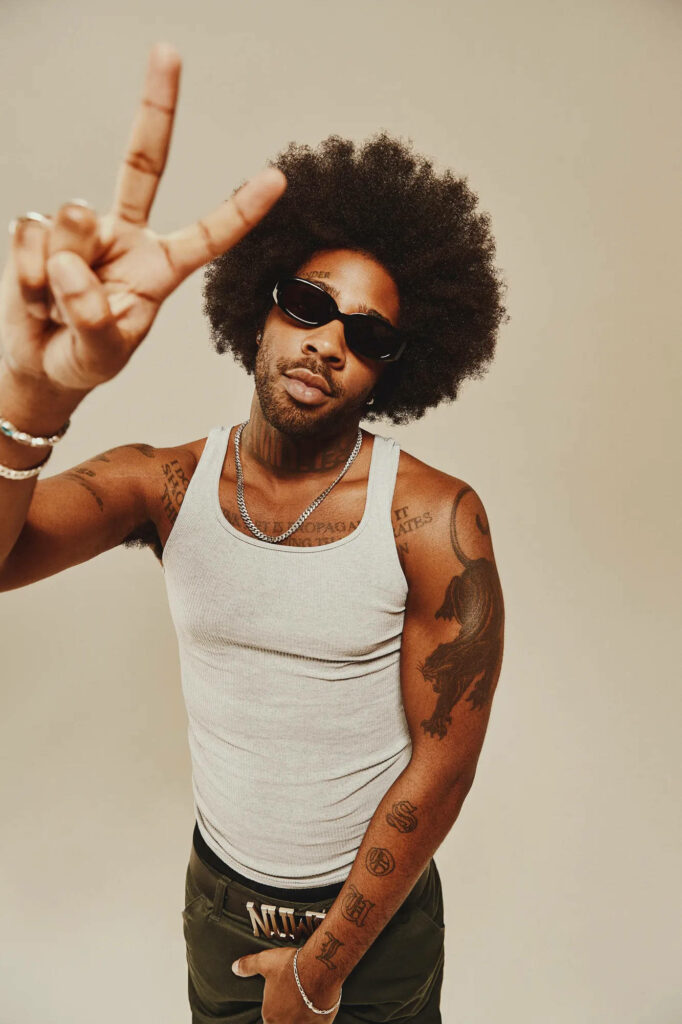 Brent Faiyaz peace sign afro wallpaper sunglasses white tank top neutral beige background