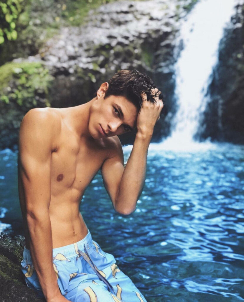 Brent Rivera shirtless by vibrant blue waterfall with lush tropical greenery summer adventure background Wallpaper