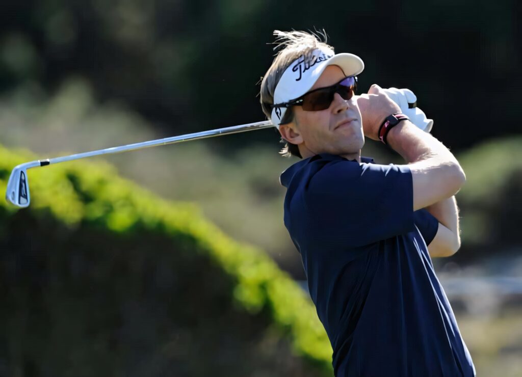 Brett Quigley mid swing golf shot wearing Titleist visor sunglasses on lush green course background Wallpaper