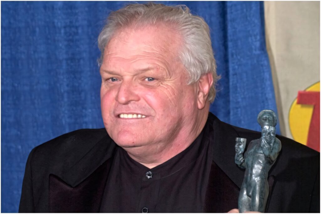 Brian Dennehy in black suit holding award trophy smiling vibrant blue gold backdrop wallpaper