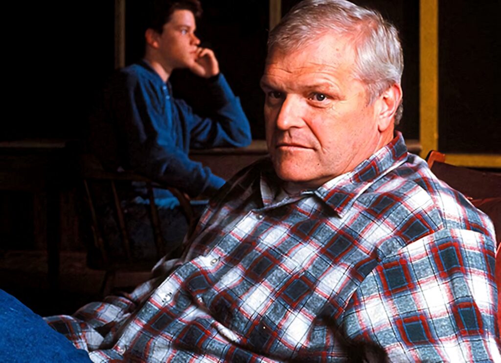 Brian Dennehy in red blue plaid flannel shirt seated indoors with intense expression rustic background Wallpaper