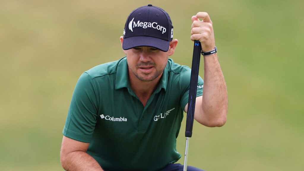 Brian Harman leaning on golf club wearing green polo and black cap on blurred golf course background Wallpaper