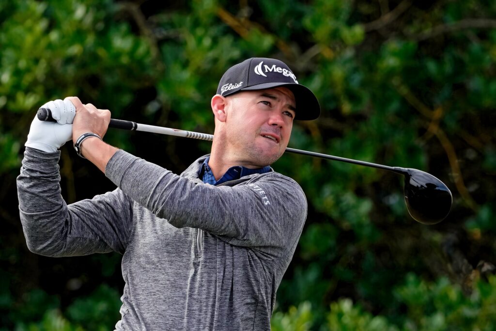 Brian Harman mid-swing in gray shirt and black cap on lush green golf course wallpaper background