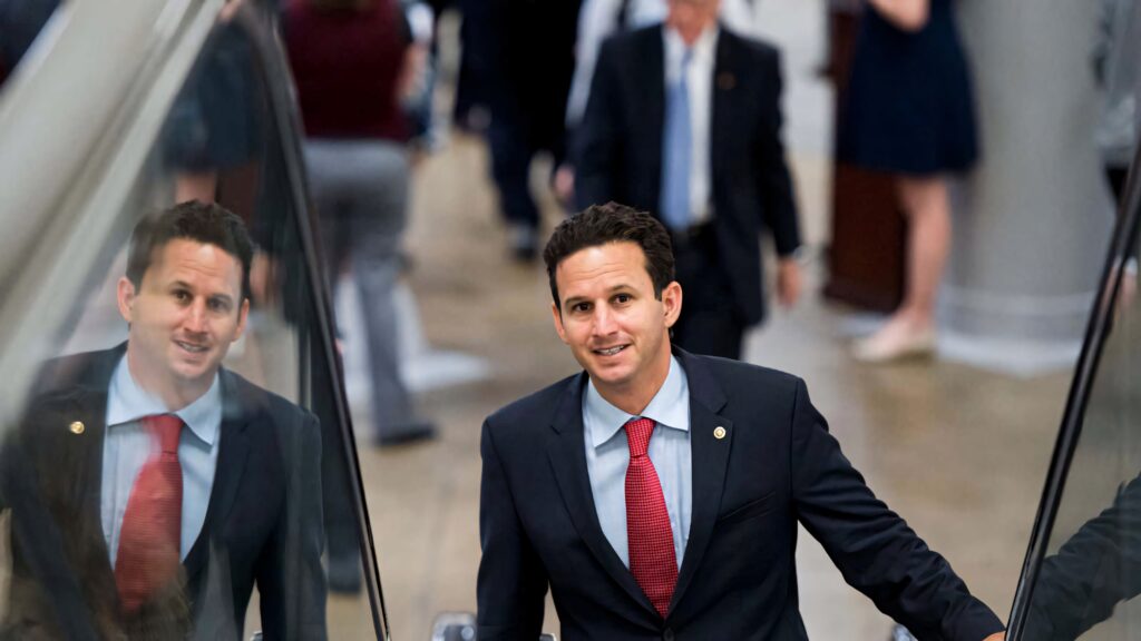 Brian Schatz in sharp suit and red tie ascending escalator with reflection and blurred crowd background Wallpaper