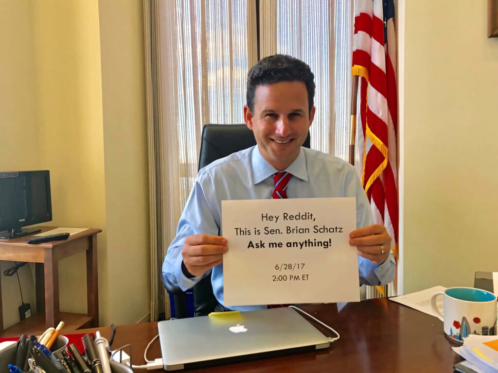 Senator Brian Schatz holding Reddit AMA sign at desk with American flag and window blinds background Wallpaper
