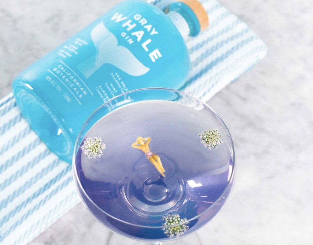 Gray Whale Gin bottle with purple cocktail garnished by flowers and swimmer on blue striped cloth Wallpaper