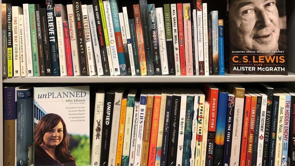Organized two-tier bookshelf Zoom background with C S Lewis A Life and unPLANNED books bright study Wallpaper