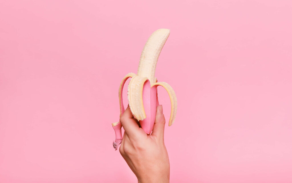 Hand holding partially peeled bright yellow banana against smooth pink wallpaper background