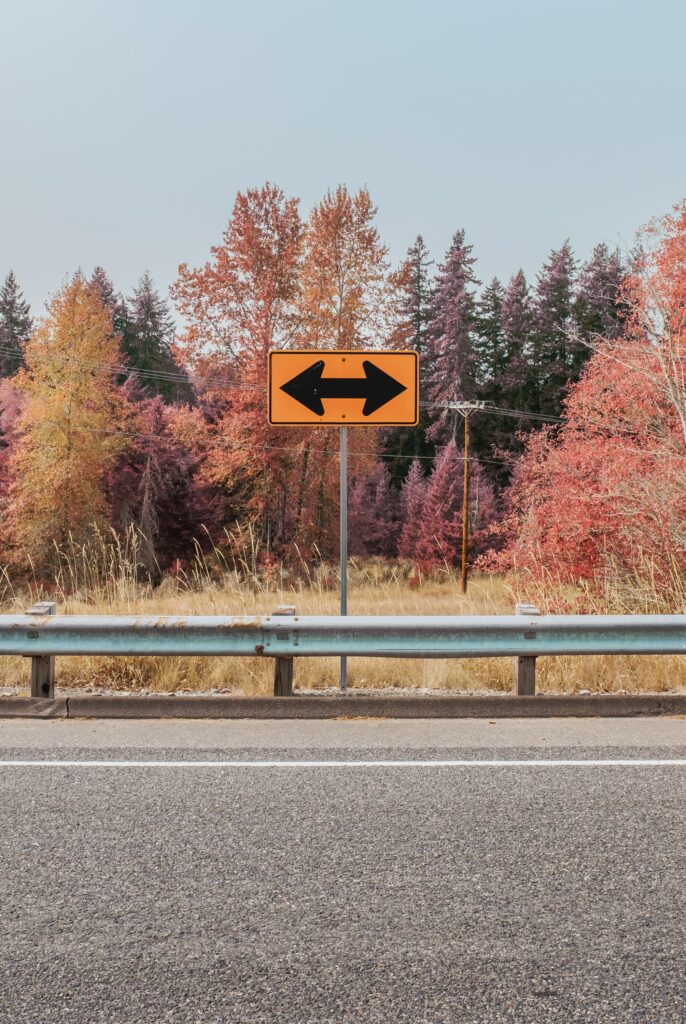 Bright yellow road sign with black arrows beside autumn trees and a quiet scenic road wallpaper