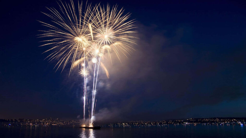 Brilliant yellow fireworks burst over calm ocean illuminating night sky with reflections wallpaper