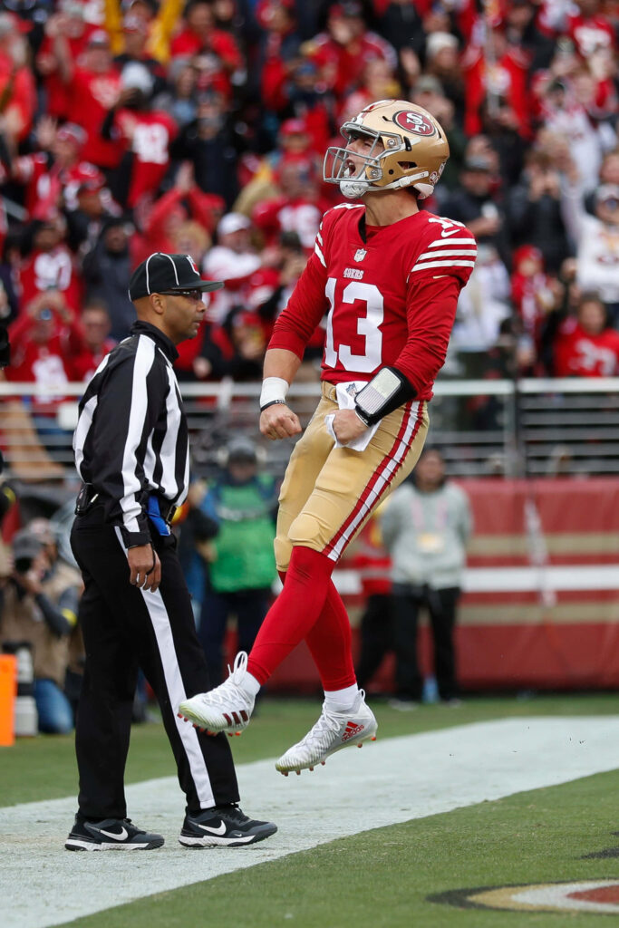 Brock Purdy 49ers mid-air touchdown celebration with referee and lively stadium crowd background Wallpaper
