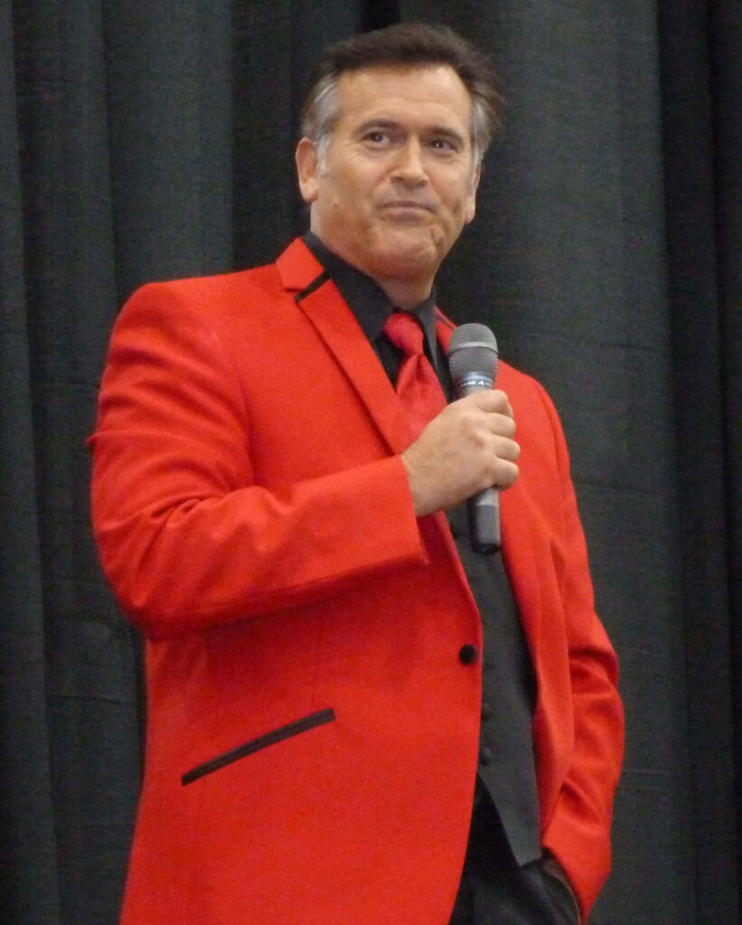 Bruce Campbell in red suit holding microphone confident pose dark background stylish wallpaper