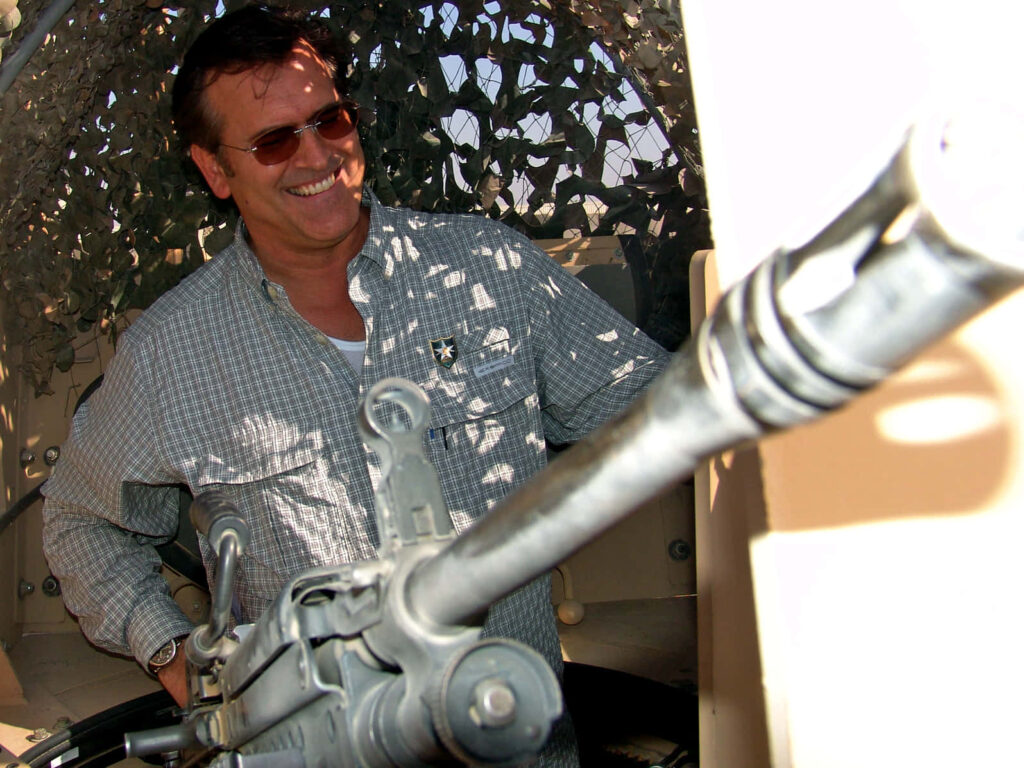 Bruce Campbell smiling in checkered shirt behind mounted gun with camo net action wallpaper