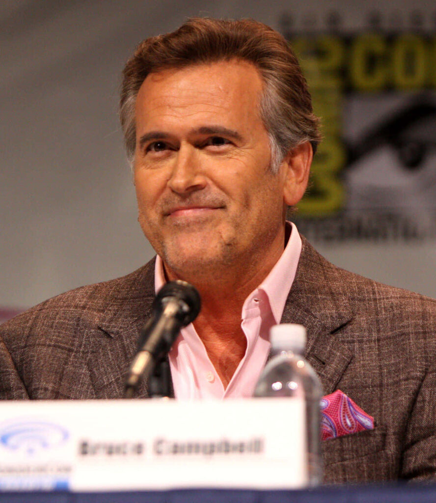 Bruce Campbell in checked brown suit smiling confidently at panel event with blurred background Wallpaper
