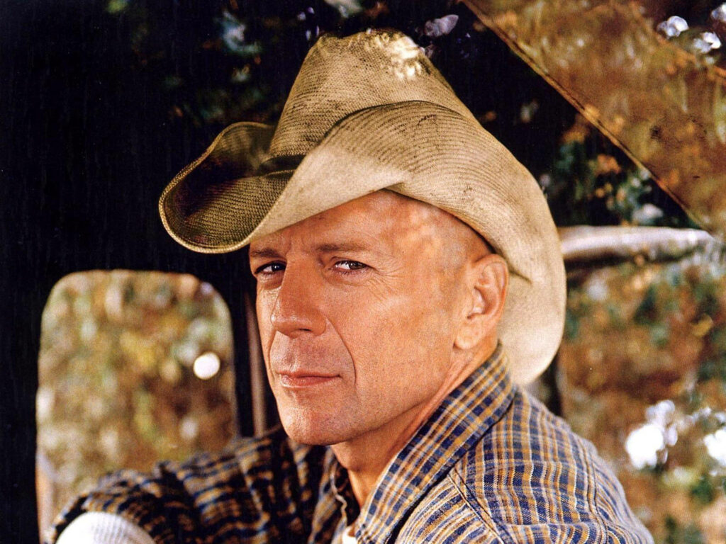 Bruce Willis as a rugged cowboy in worn hat and plaid shirt against rustic outdoor background Wallpaper