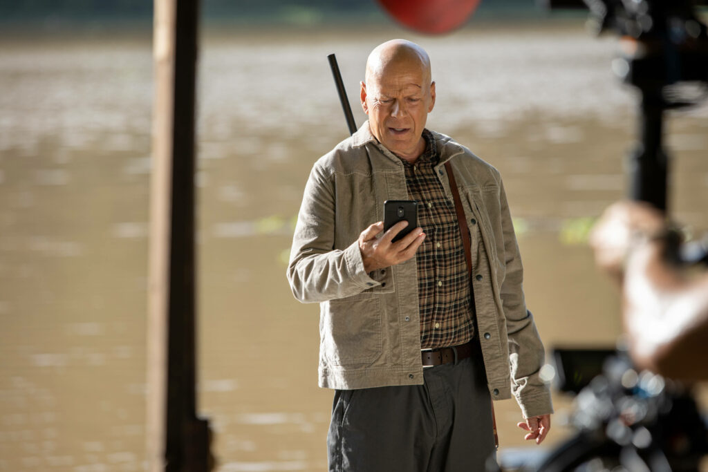 Bruce Willis in rugged action outfit by lake checking phone outdoors cool adventurous wallpaper