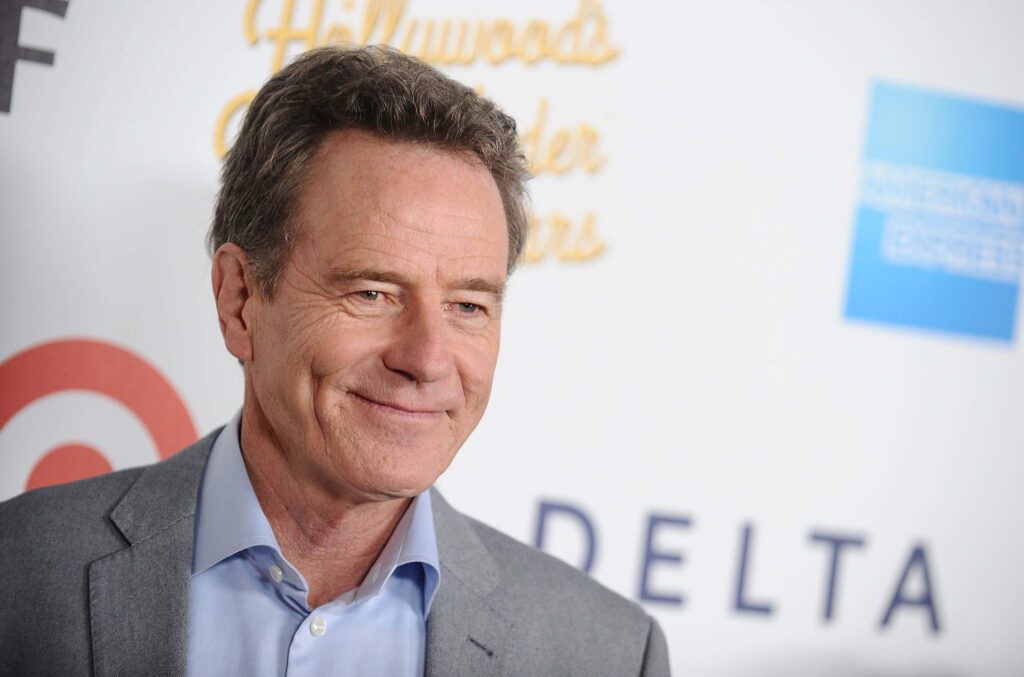 Bryan Cranston in grey suit smiling confidently with white and gold event backdrop wallpaper
