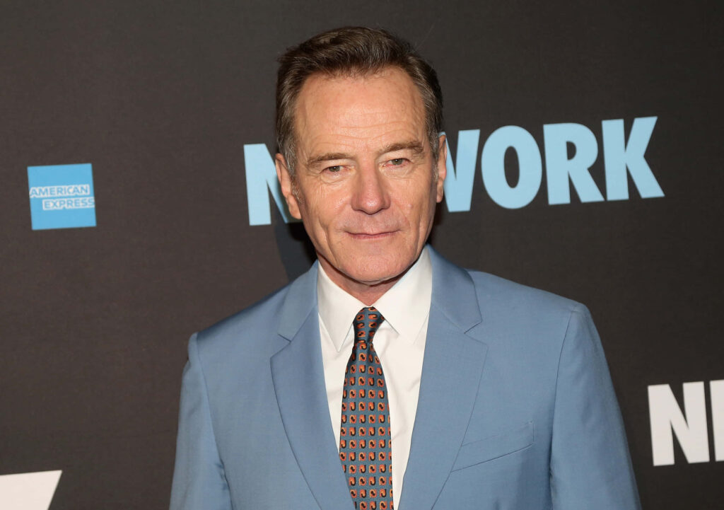 Bryan Cranston in sharp light blue suit with patterned tie on bold black and blue wallpaper background
