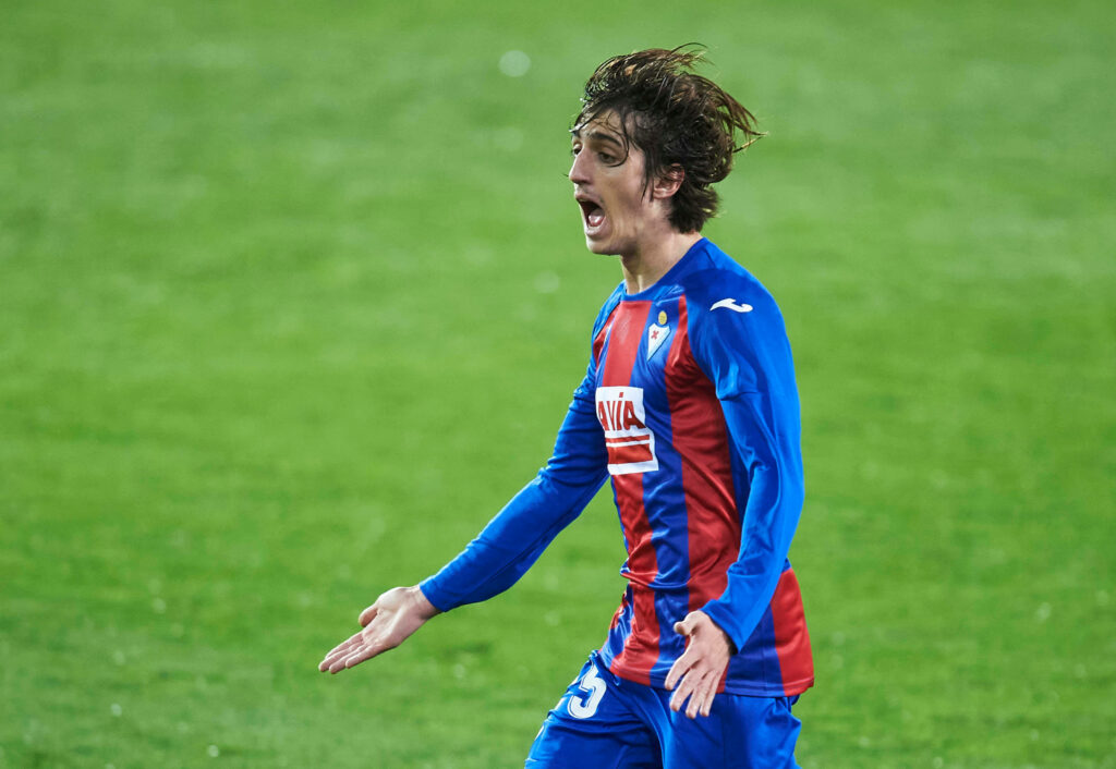 Bryan Gil intense football action wallpaper in red and blue Eibar jersey on vibrant green field