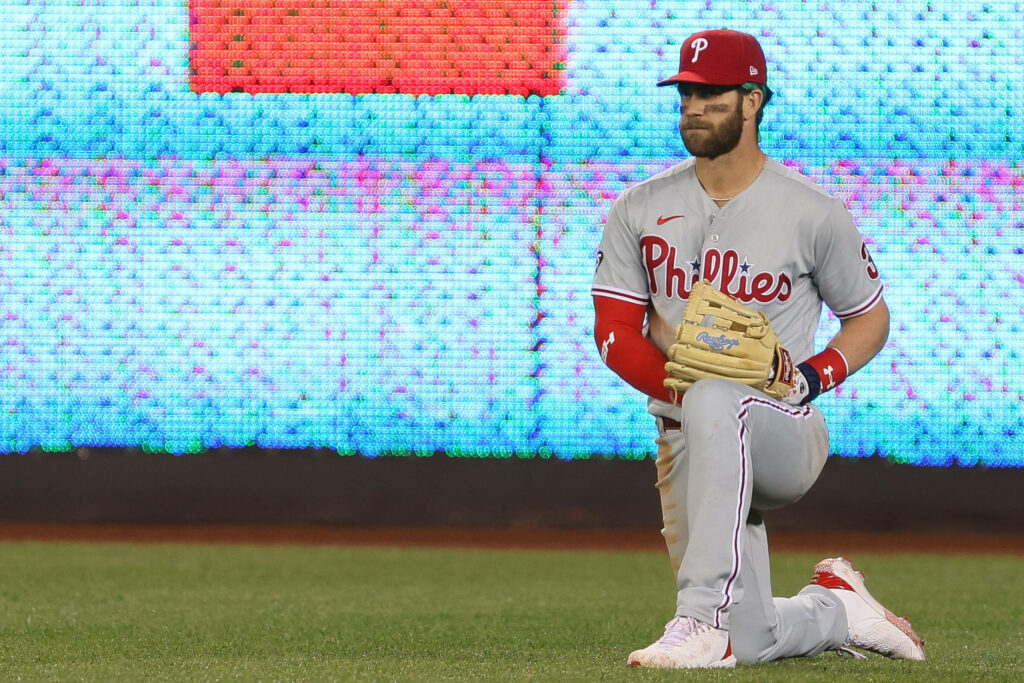 Bryce Harper Phillies baseball gloves wallpaper kneeling on field with vibrant scoreboard background