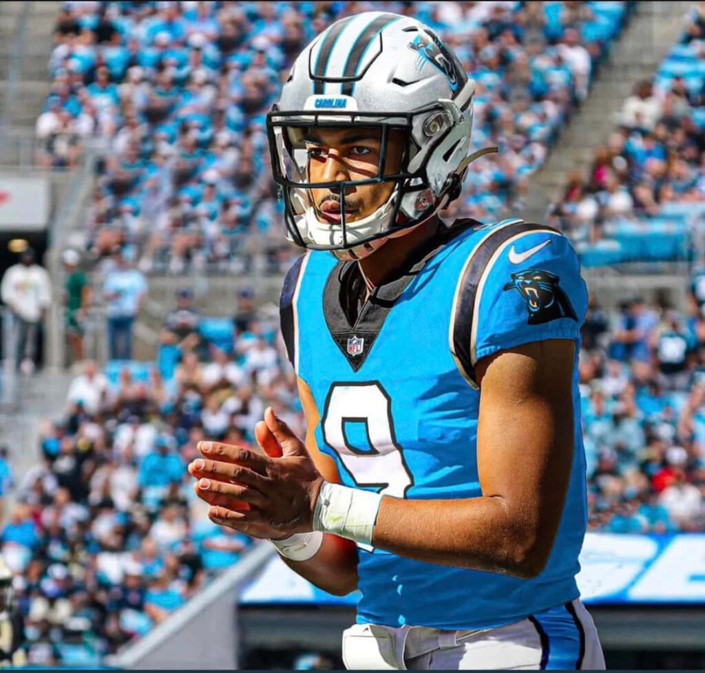 Bryce Young in blue Carolina Panthers jersey focused on field with vibrant stadium crowd background Wallpaper