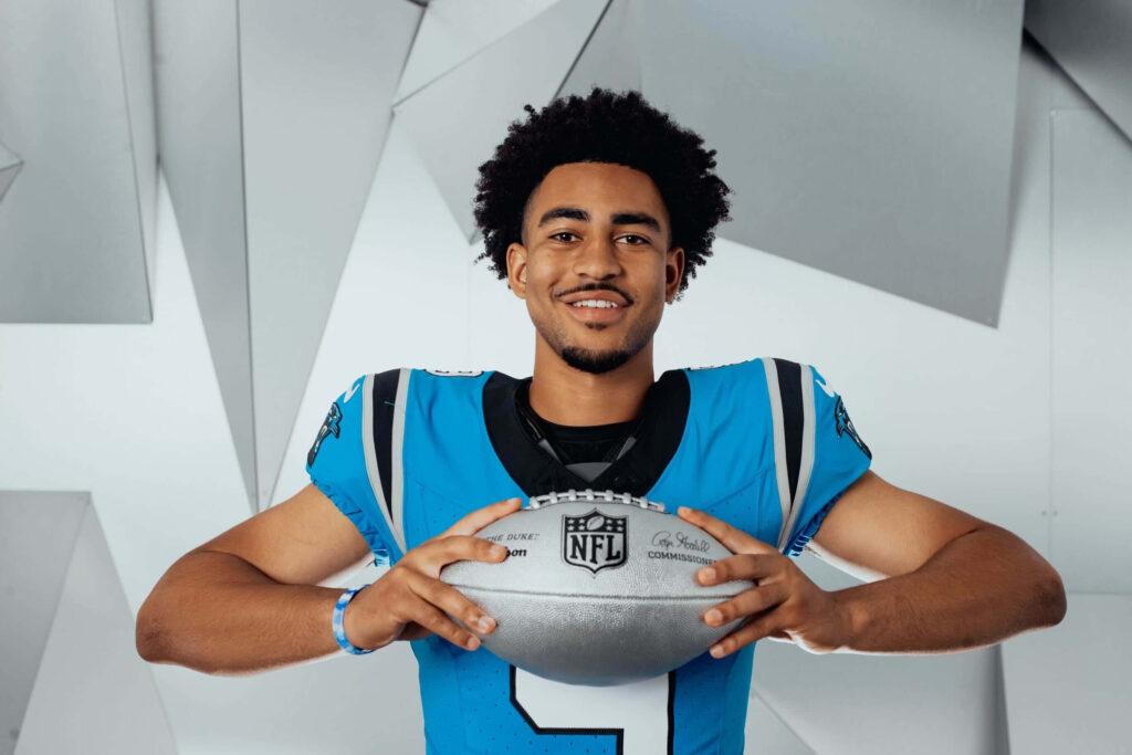 Bryce Young in Carolina Panthers uniform holding NFL football smiling dynamic geometric background Wallpaper