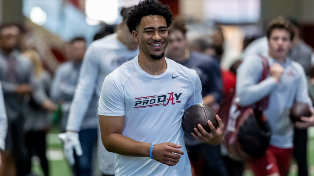Bryce Young smiling confidently holding football at pro day with athletes and trainers background Wallpaper