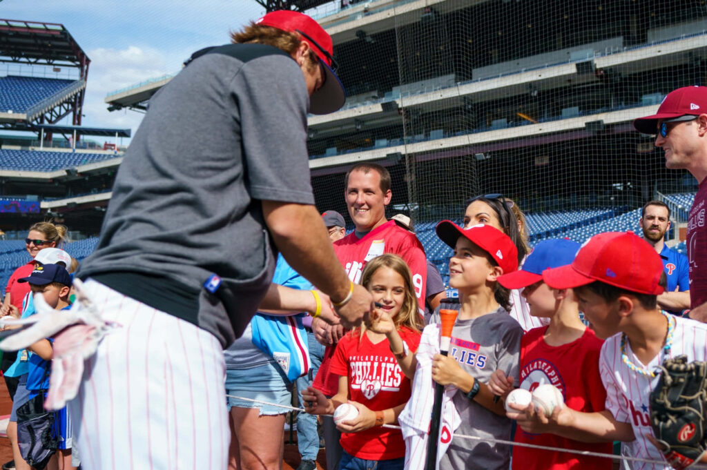 Bryson Stott signing autographs for young Phillies fans at sunny ballpark with excited crowd Wallpaper
