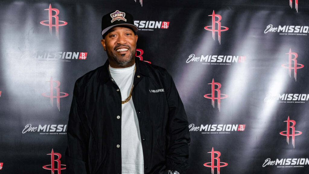 Bun B in UGK cap black jacket gold chain poses at Houston Rockets One Mission banner event Wallpaper