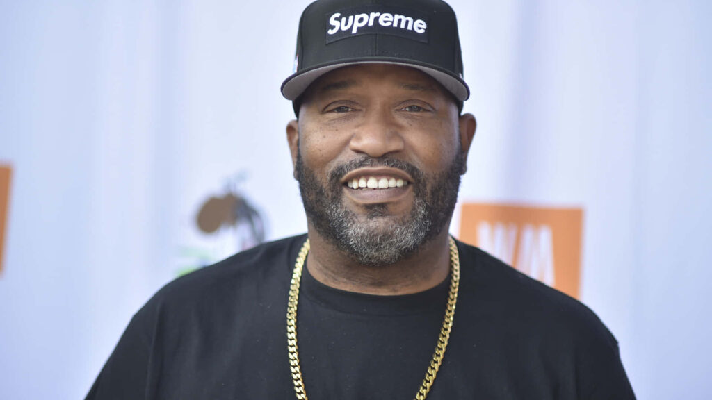 Bun B wearing black Supreme cap and shirt with gold chain smiling in realistic wallpaper background
