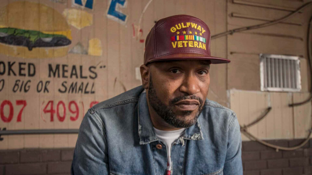 Bun B wearing maroon Gulfway Veteran cap denim jacket against rustic mural wall wallpaper
