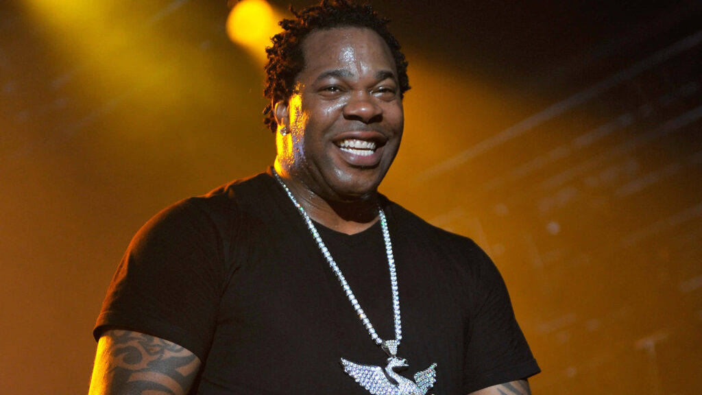 Busta Rhymes performing on stage under yellow lights wearing black t-shirt and statement chain Wallpaper