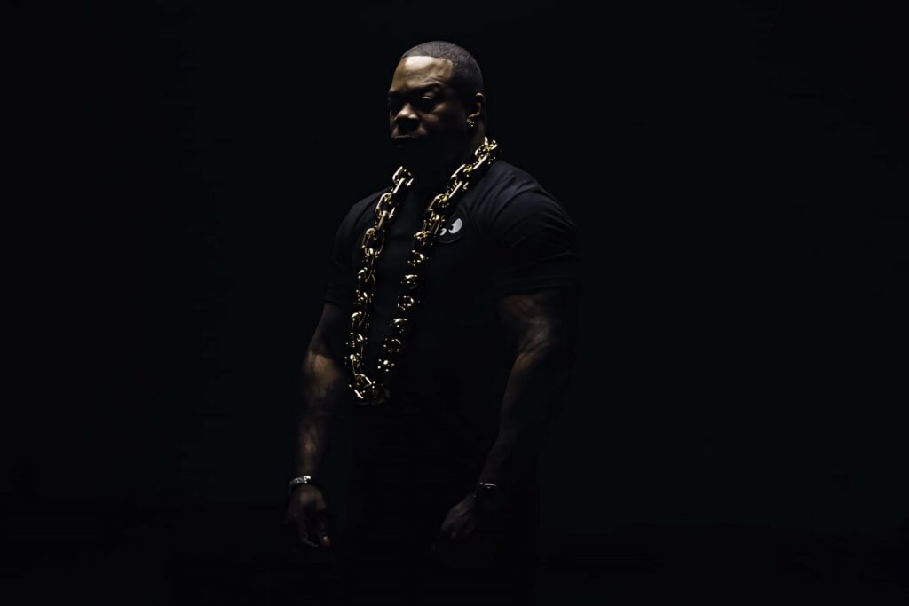 Busta Rhymes in low-key lighting with gold chain and muscular build on dark minimalist wallpaper