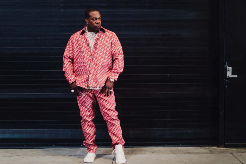 Busta Rhymes in bold red patterned outfit with white sneakers posing against dark industrial backdrop Wallpaper