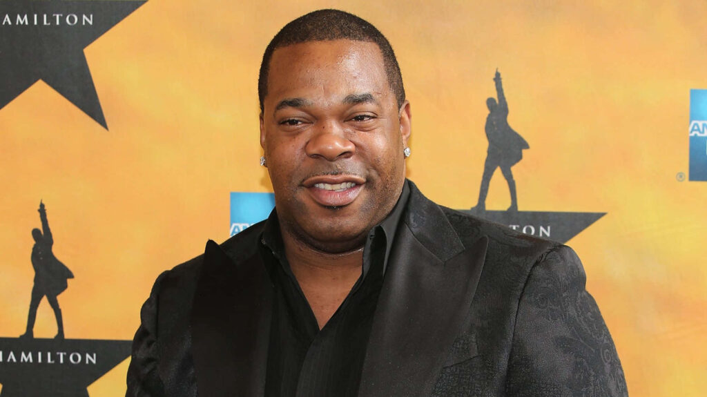 Busta Rhymes in black suit smiling at award event with bright yellow Hamilton musical backdrop Wallpaper
