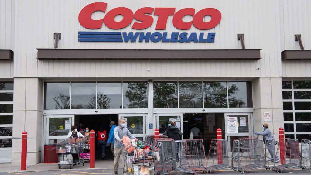 Shoppers with carts outside busy Costco Wholesale store under large red blue logo wallpaper background