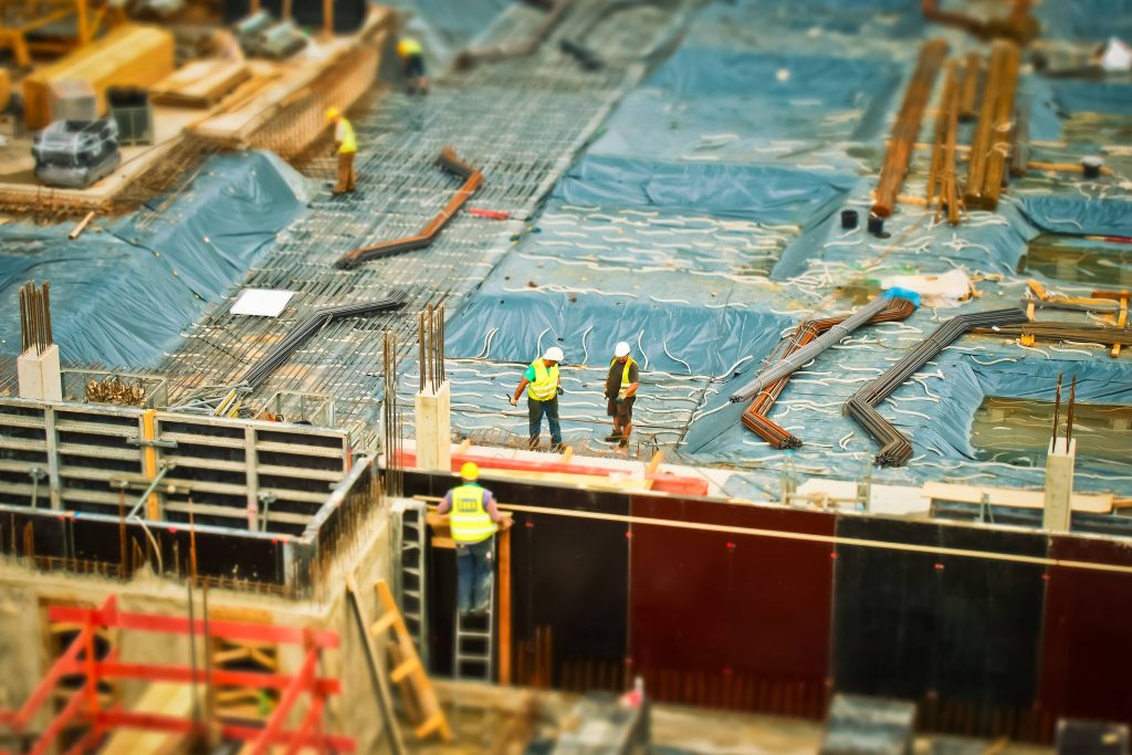 Construction workers in bright gear at busy site with rebar bundles and blue tarps tilt-shift Wallpaper