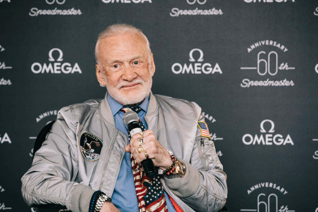 Buzz Aldrin in silver jacket with mission patches and American flag against Omega Speedmaster background Wallpaper