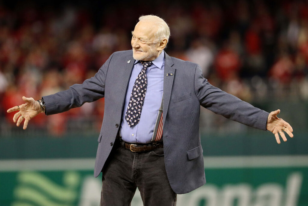 Buzz Aldrin speaking on stage in blue blazer and patterned tie with vibrant stadium background Wallpaper